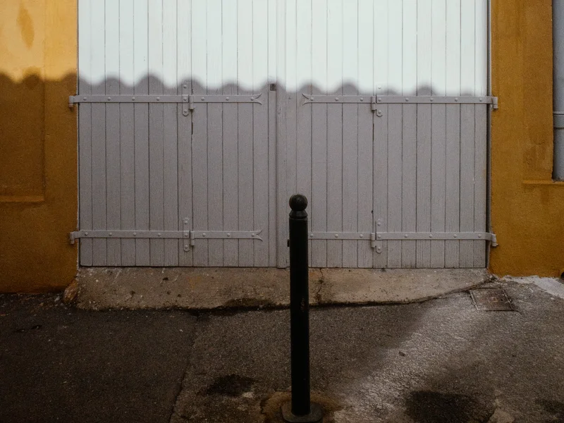 A white wooden door set in a yellow wall with a shadow and a black bollard in front.