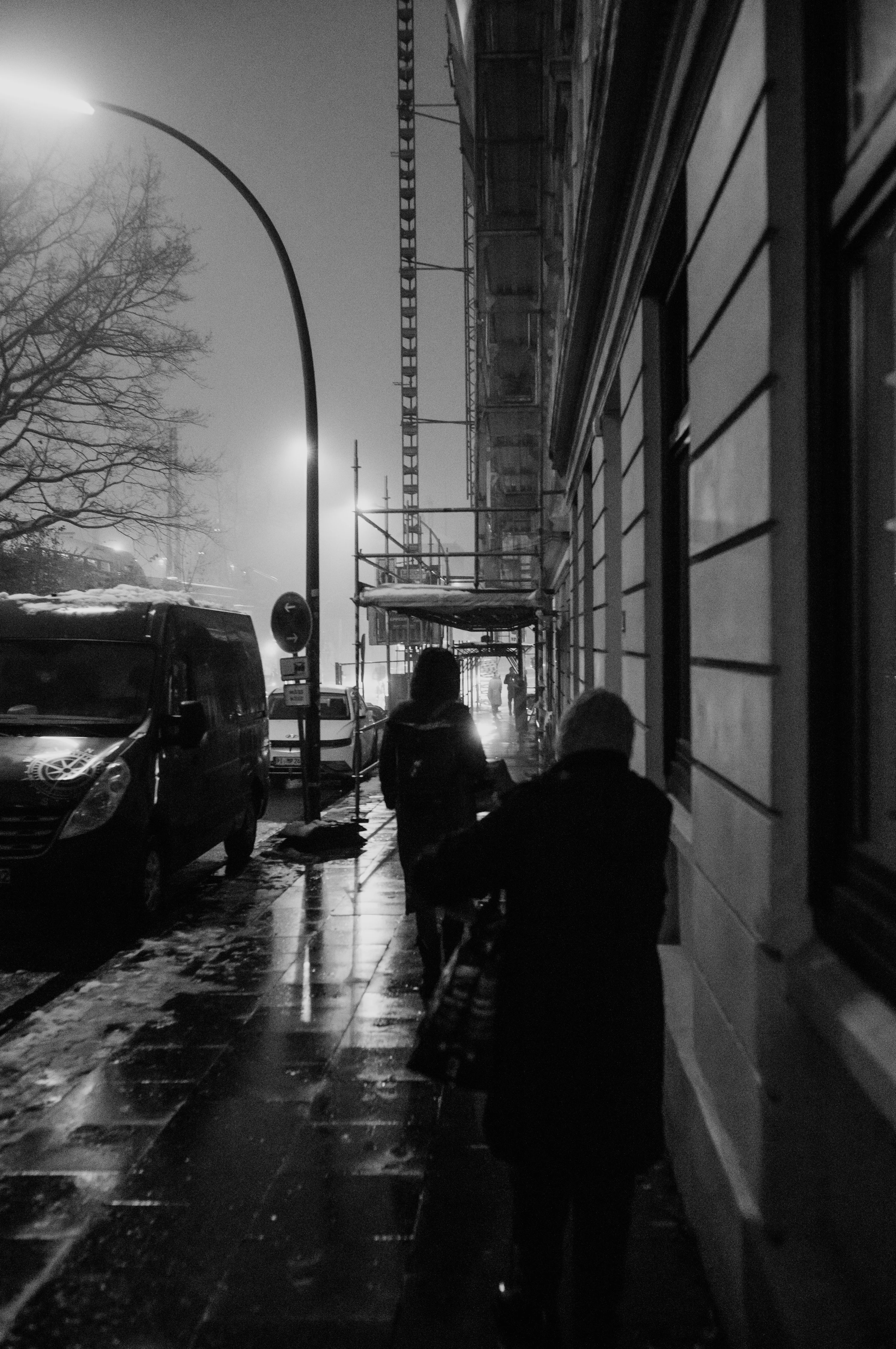 Two people walking on a wet, dimly lit street at night with buildings and cars lining the side.