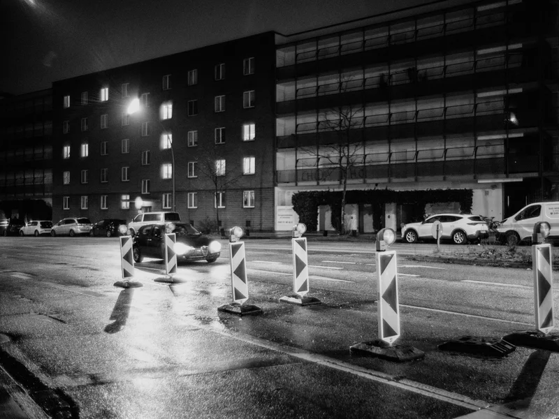A nighttime street scene featuring a car driving past road barriers in front of a dimly lit building.