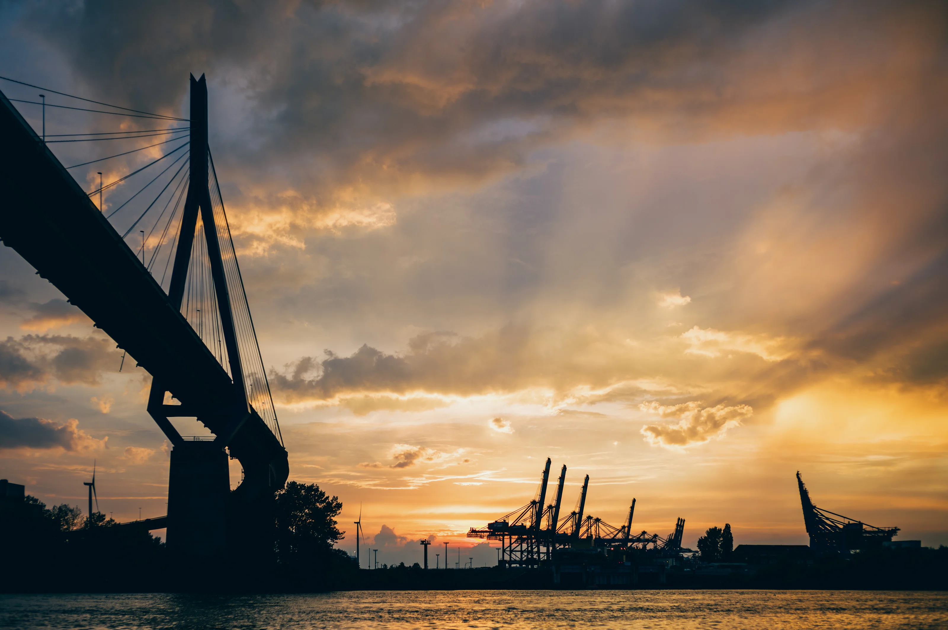 Sunset view of a bridge and port cranes silhouetted against a cloudy sky.