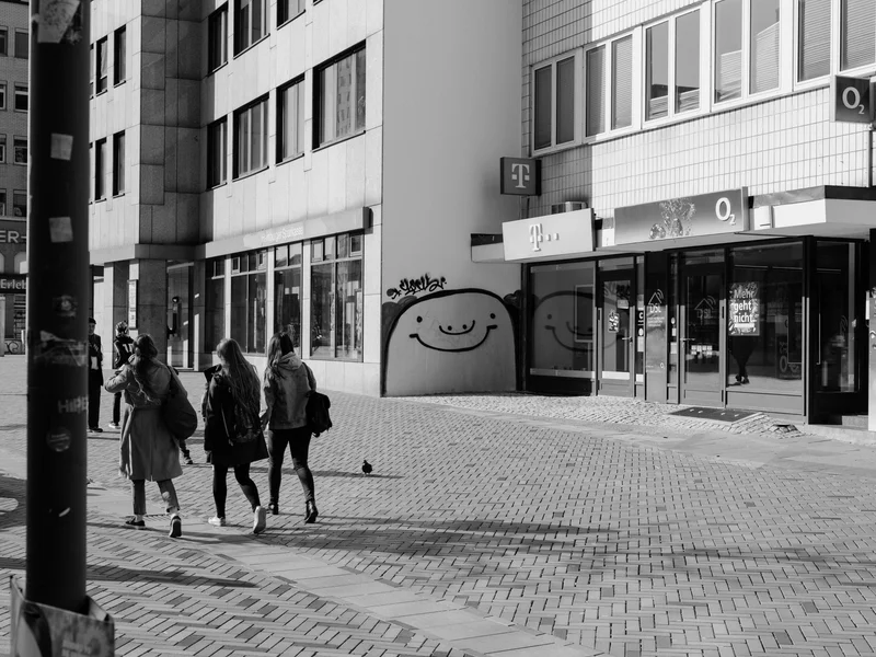 A group of people walking past a building with a smiling graffiti face on the wall.
