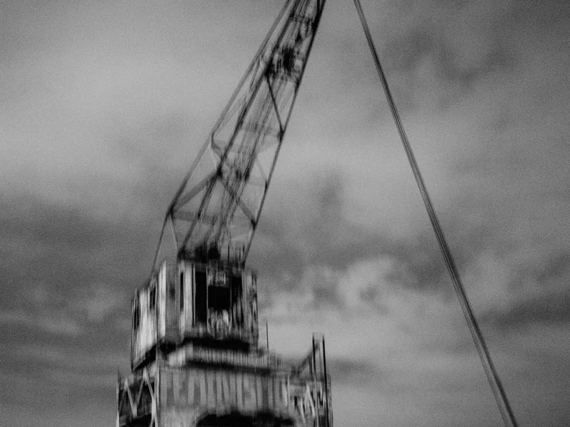 Blurry black and white image of an industrial crane by a waterfront at night.