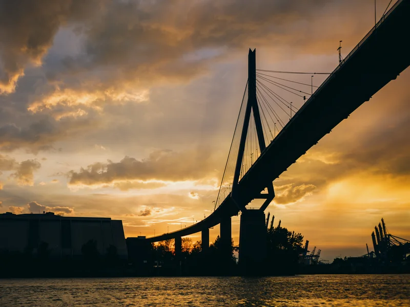 Silhouette of a suspension bridge at sunset with dramatic clouds and orange sky.