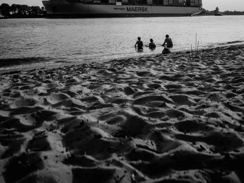 Three people sitting on a sandy beach with a large container ship passing by on the water.