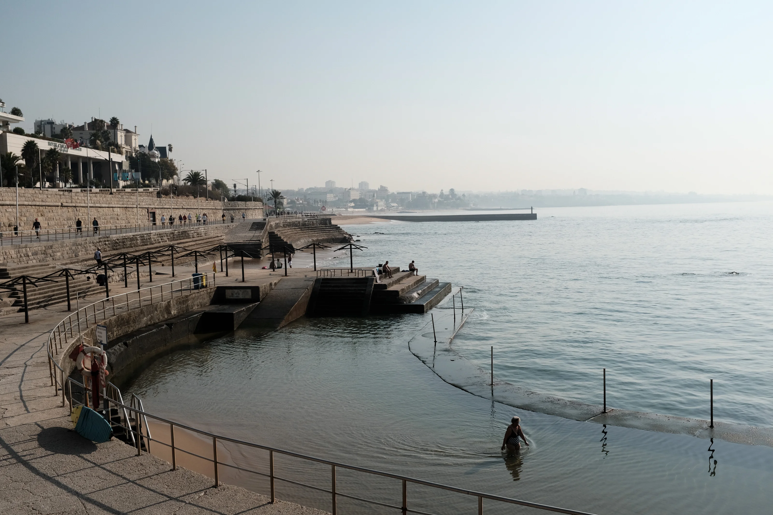 Seaside promenade with people walking and a person wading in the water near steps and railings.