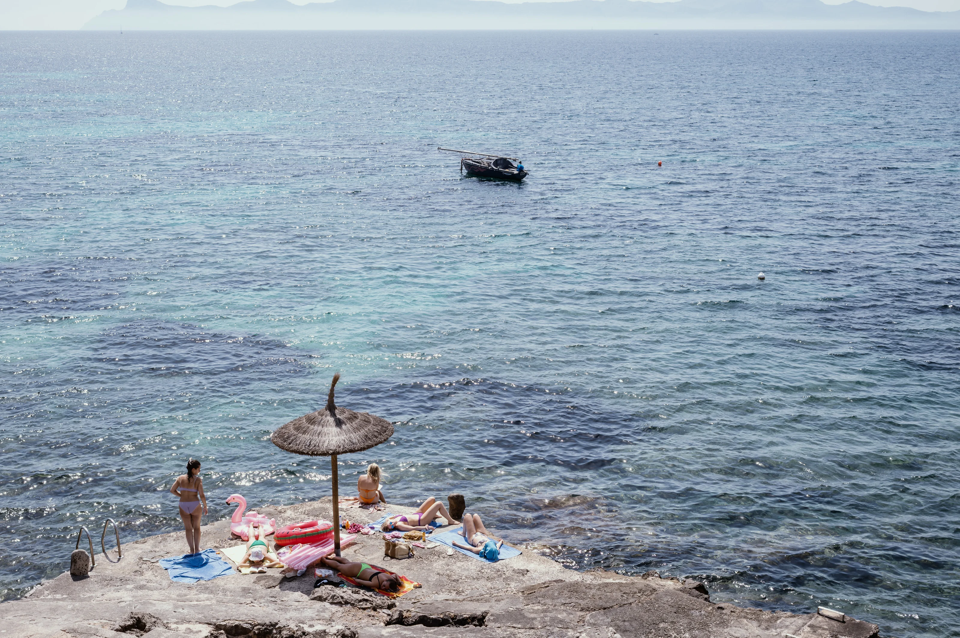 People relaxing with inflatables on a rocky beach by the sea under a straw parasol.