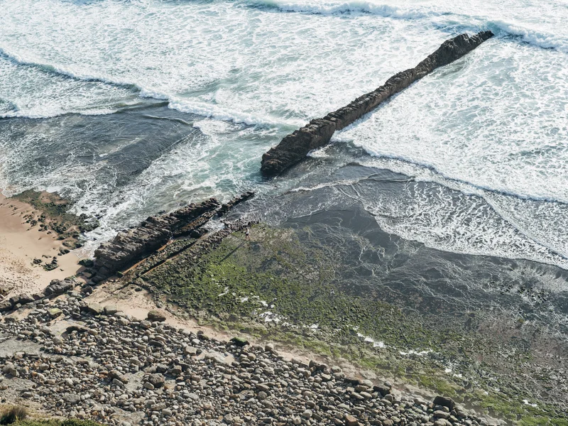 Coastal scene with waves crashing against a rocky shore and a long rock formation extending into the ocean.