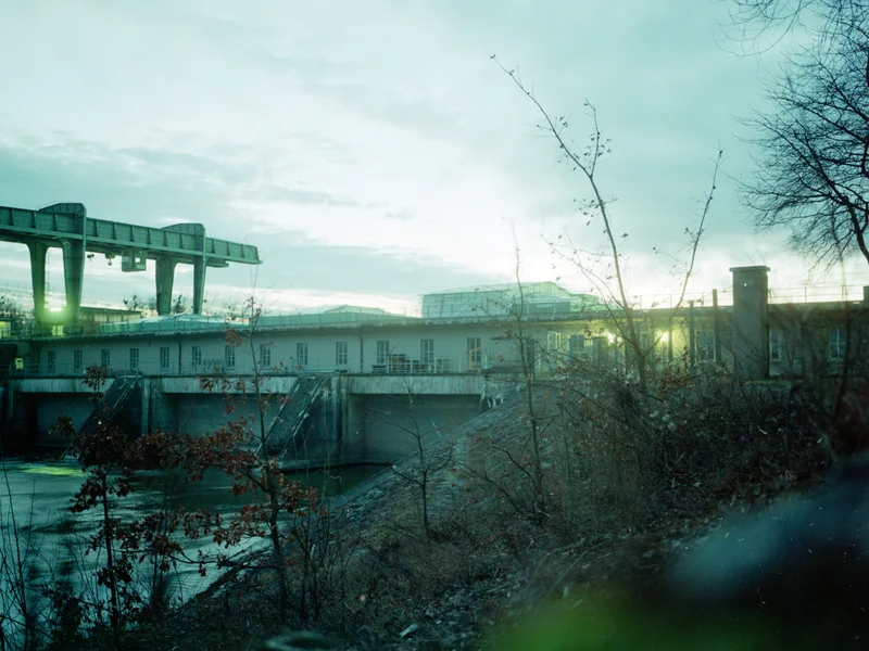 Industrial building and structures silhouetted against a cloudy sky with bare trees in the foreground.