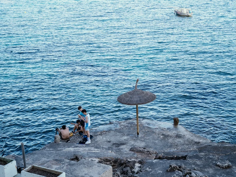 Group of people on a rocky shore next to a straw umbrella and calm blue sea.