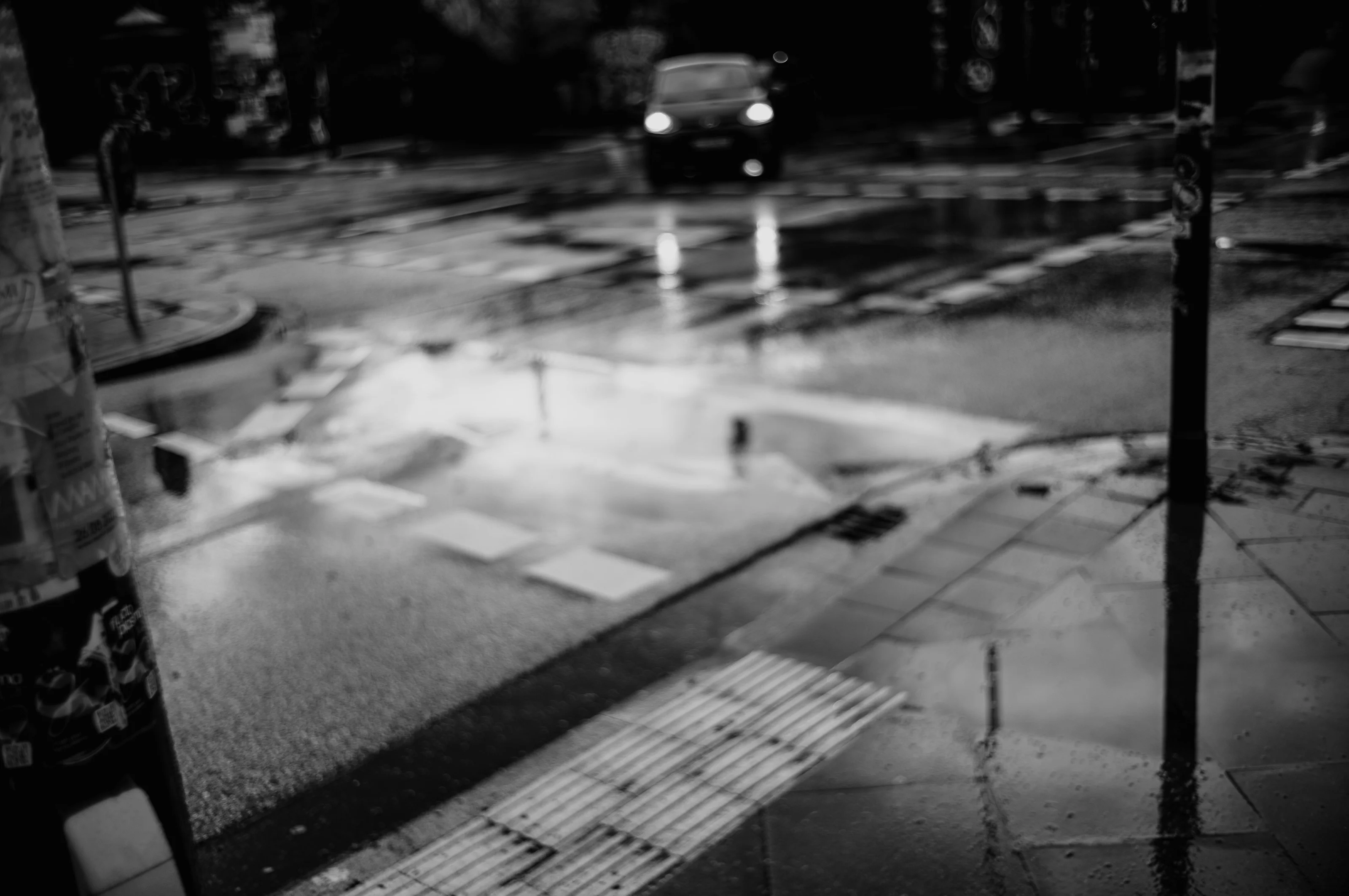 A blurry black and white street scene with a car approaching and reflections on wet pavement.