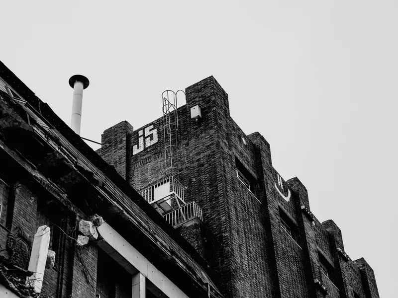 Black and white photo of an old industrial building with a chimney and 'J15' marked on the brick wall.