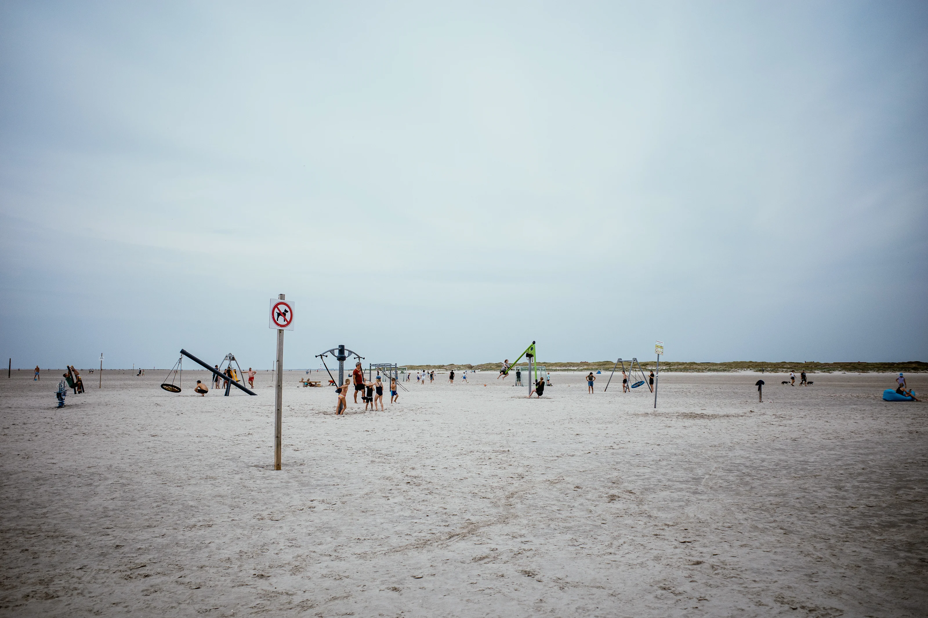A beach scene featuring a playground with people scattered across the sandy area under a cloudy sky.