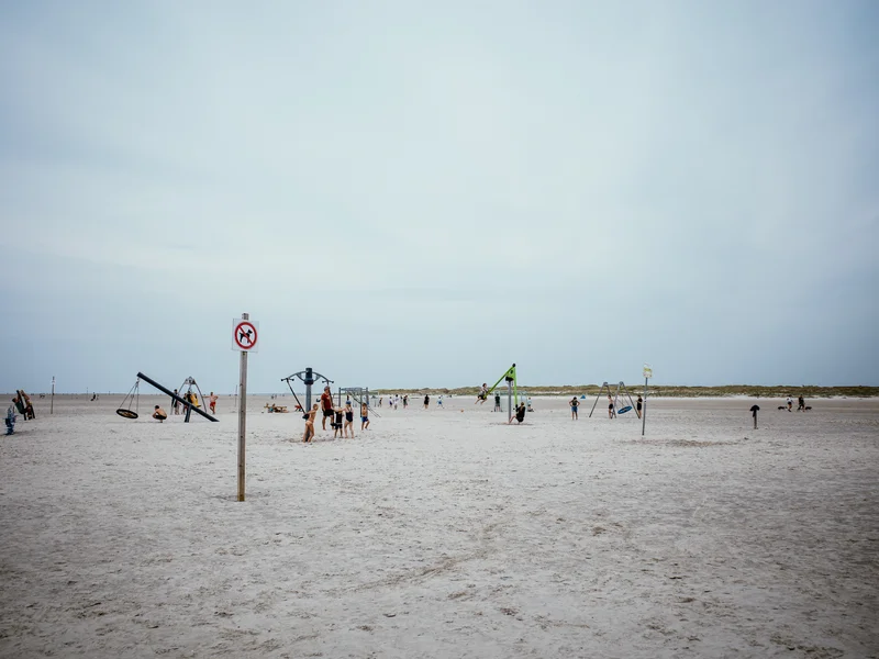 A beach scene featuring a playground with people scattered across the sandy area under a cloudy sky.