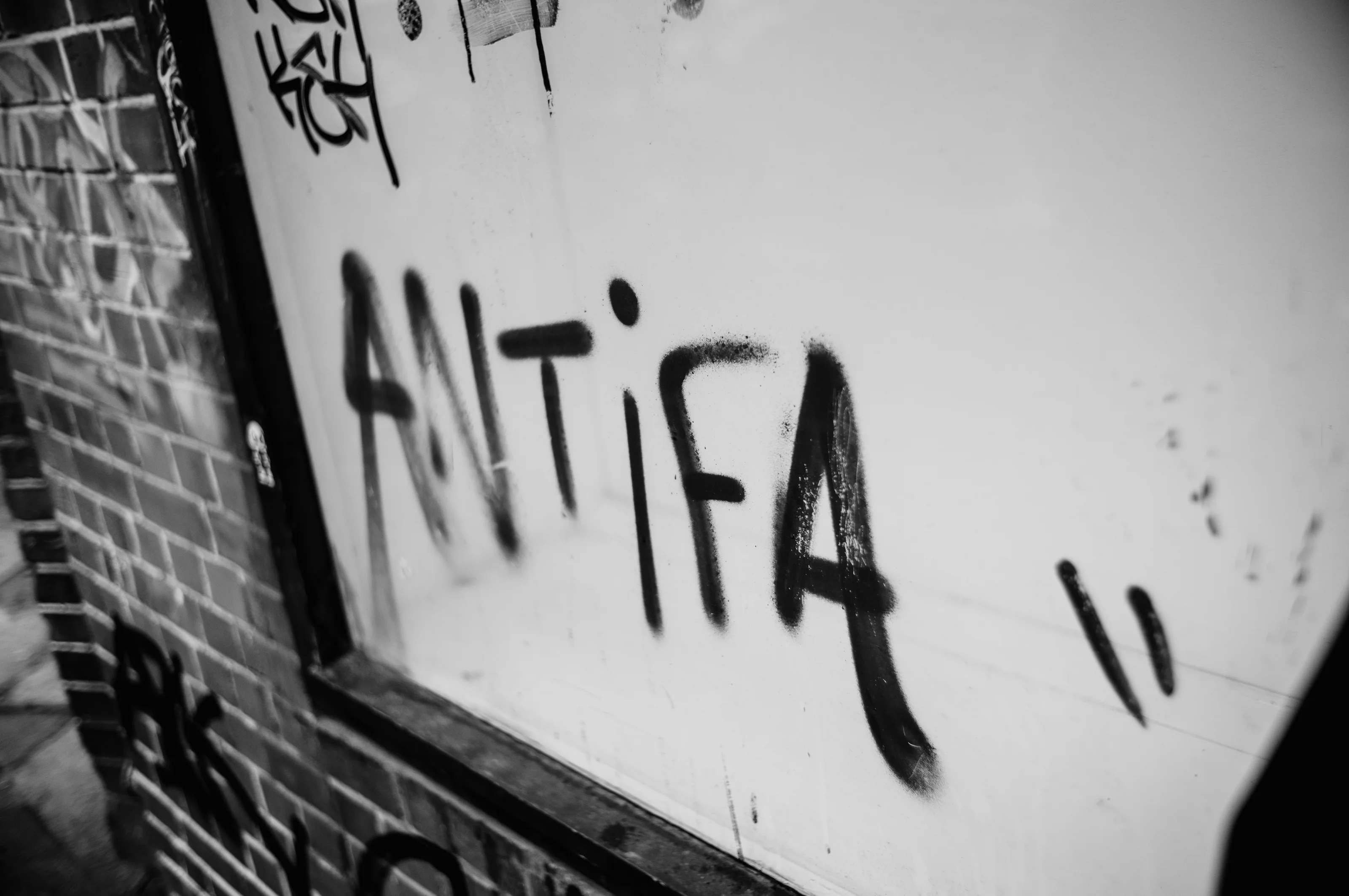 Black graffiti reading 'ANTIFA' on a white wall next to a brick surface.