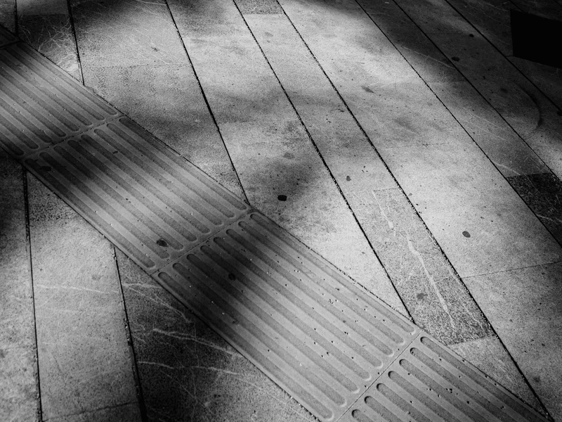 A black and white image of a textured sidewalk with shadows and a tactile paving strip.