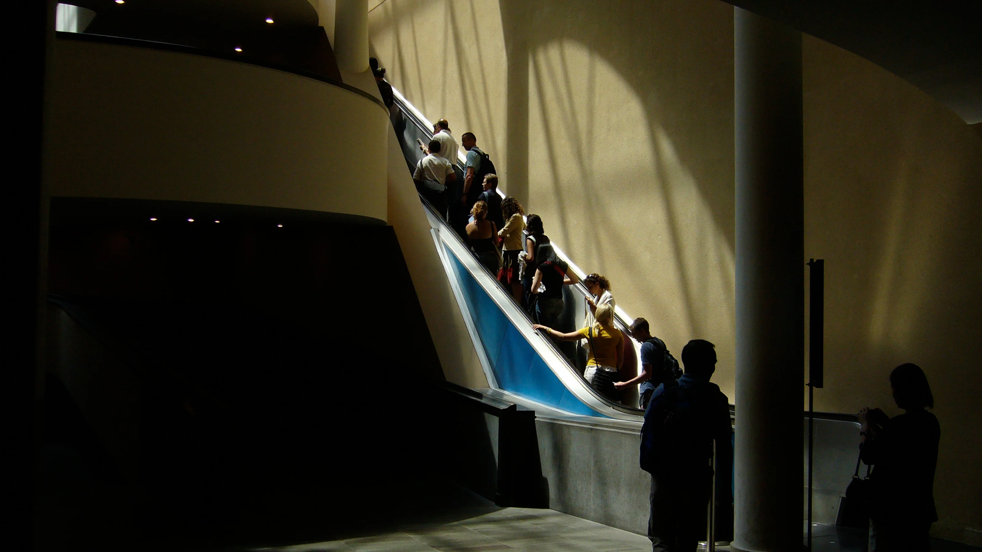 People riding an upward escalator in a building with sunlight casting shadows.