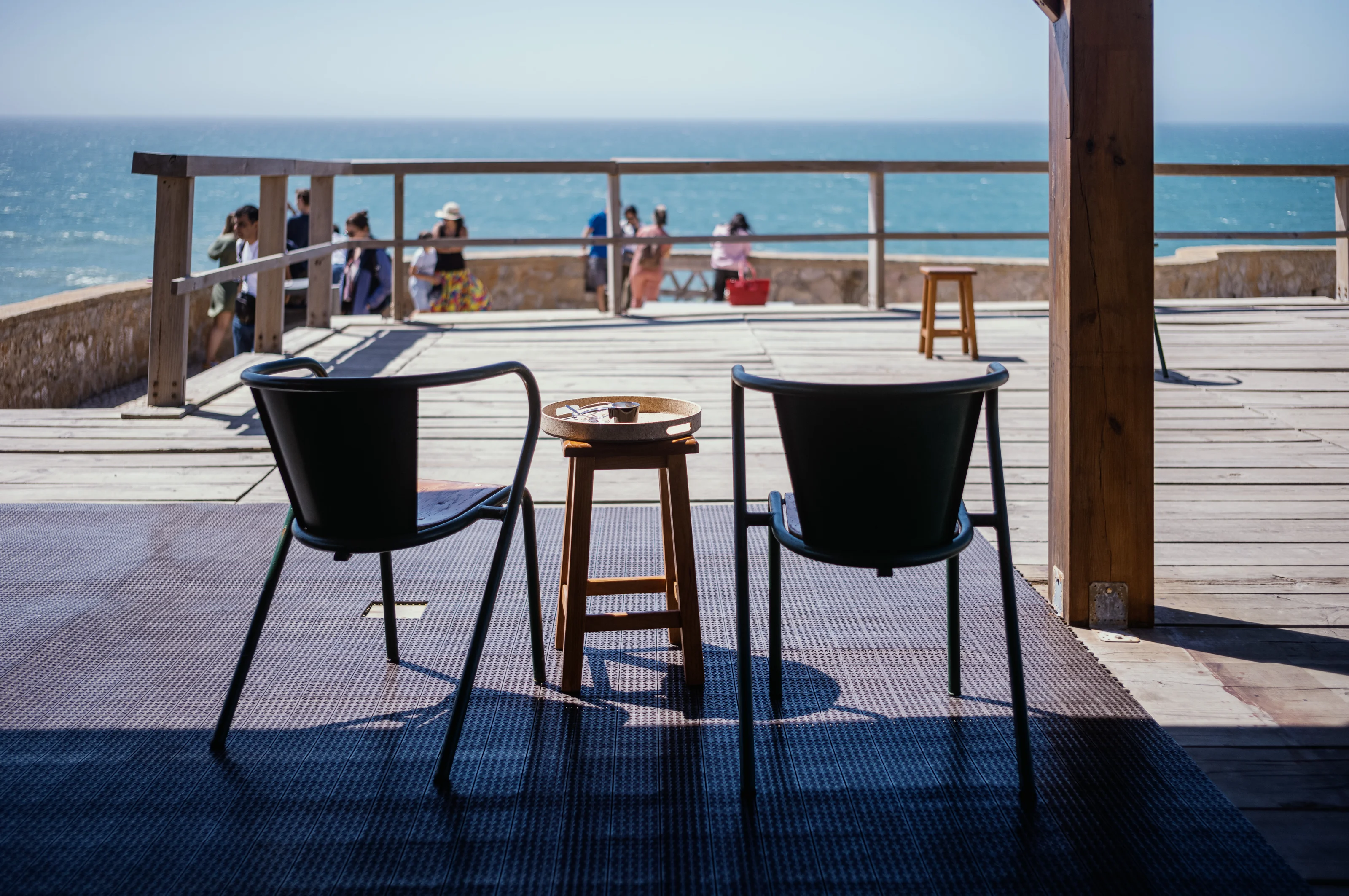 Two chairs and a small table facing the sea with people in the background on a deck.