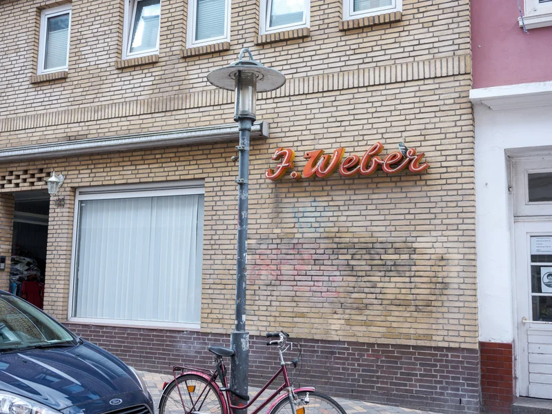 A brick building facade with the name 'F. Weber' in neon letters above a bicycle and a street lamp.