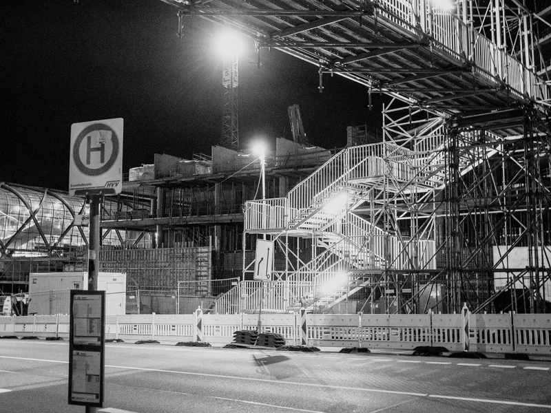A night scene showing a construction site with illuminated scaffolding and an overpass.